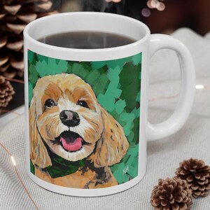 May include: White ceramic mug featuring a colorful illustration of a happy, light brown dog with a green background. The dog has a pink tongue and is wearing a green scarf. The mug is filled with a dark liquid.