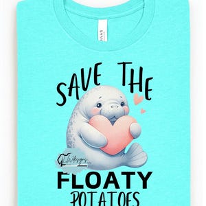 Puede incluir: Camiseta azul claro con un dibujo de manatí sosteniendo un corazón rosa. El texto "SAVE THE FLOATY POTATOES" está impreso encima y debajo de la ilustración del manatí. La camiseta está doblada y tiene una etiqueta.