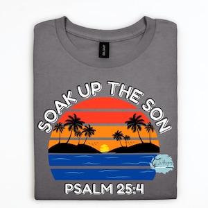 Puede incluir: Camiseta gris con un gráfico de puesta de sol con palmeras, olas del océano y el texto "SOAK UP THE SON" y "PSALM 25:4". El diseño incluye colores naranja, rojo, amarillo, azul y negro.