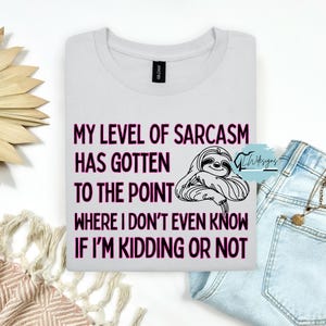 Può includere: T-shirt bianco con la scritta "MY LEVEL OF SARCASM HAS GOTTEN TO THE POINT WHERE I DON'T EVEN KNOW IF I'M KIDDING OR NOT" in rosa e un'illustrazione di bradipo nero. La maglietta è piegata su una superficie bianca.