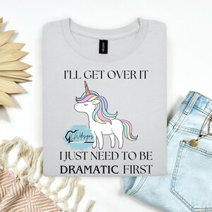 May include: White t-shirt with a unicorn graphic and the text "I'll Get Over It, I Just Need To Be Dramatic First." The unicorn has a rainbow mane and horn. The shirt is folded.