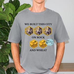 Op de afbeelding: Grijze t-shirt met de tekst "WE BUILT THIS CITY ON ROCK AND WHEAT". Het ontwerp bevat drie zeshoekige berg afbeeldingen en twee tarwe afbeeldingen. Het shirt is een casual stijl.