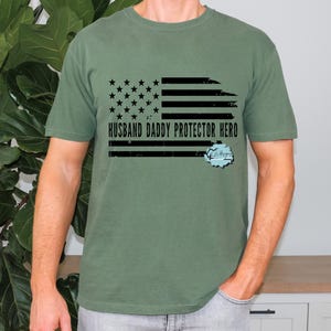 May include: Olive green t-shirt featuring a distressed American flag design with the text "HUSBAND DADDY PROTECTOR HERO". The graphic includes stars, stripes, and a light blue and white design.