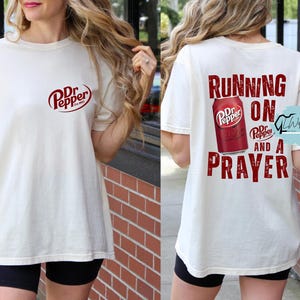 May include: Cream-colored t-shirt featuring the Dr. Pepper logo on the front and a graphic of a Dr. Pepper can with the words "Running on and a Prayer" on the back. The shirt is a casual, short-sleeved style.
