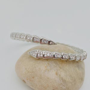 Può includere: Un bracciale aperto color argento con un design segmentato che ricorda un serpente. La superficie metallica è incastonata con numerose piccole pietre chiare e scintillanti disposte in un motivo ripetitivo lungo tutta la fascia flessibile.