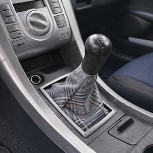 May include: Close-up of a car's interior, focusing on the gear stick. The black gear knob is paired with a plaid fabric boot. The console is silver and black. A blue patterned seat is visible in the background.
