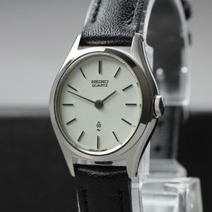 Könnte beinhalten: Eine silberne Seiko Quartz Armbanduhr mit weißem Zifferblatt und schwarzen Zeigern. Die Uhr hat ein schwarzes Lederarmband. Das Zifferblatt hat schwarze Markierungen und das Seiko Quartz Logo. Die Uhr wird auf einem transparenten Ständer ausgestellt.