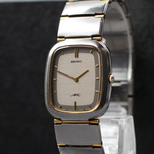 Könnte beinhalten: Eine Vintage Seiko Arc Armbanduhr mit einem silber- und goldfarbenen rechteckigen Zifferblatt und einem silbernen Armband. Das Zifferblatt zeigt das Seiko-Logo und das Wort "ARC" darunter. Die Uhr hat goldfarbene Zeiger und Stundenmarkierungen.