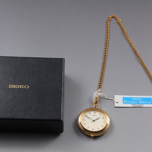 May include: A gold-toned Seiko pocket watch with a cream-colored face and a matching chain, presented with a black box. The watch has a tag that reads "SEIKO POCKET WATCH".