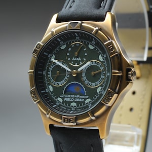 May include: A gold-toned ALBA Field Gear watch with a dark green face and black leather band. The watch face displays multiple subdials and the text "WATER 10 BAR RESIST". The watch is on a light gray background.