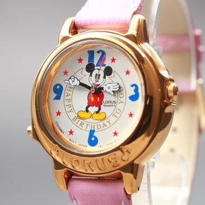 Pode incluir: Um relógio Lorus Quartz dourado com uma pulseira de couro rosa. O mostrador apresenta Mickey Mouse, algarismos azuis e o texto "Happy Birthday to You".