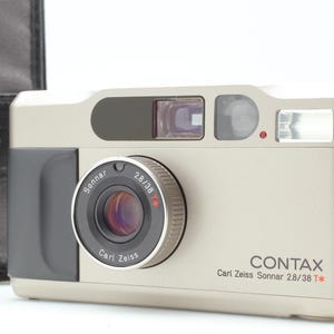 Peut inclure: Un appareil photo argentique Contax T2 couleur champagne avec une poignée noire et un objectif Carl Zeiss Sonnar 2.8/38 T*. L'appareil présente un boîtier rectangulaire, un flash intégré, un viseur et une fenêtre de télémètre. Étui noir inclus.