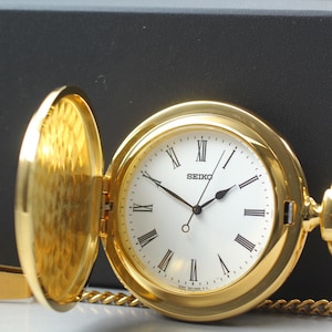 Puede incluir: Un reloj de bolsillo Seiko dorado con esfera blanca y números romanos negros. El reloj está abierto, revelando la esfera y el interior texturizado de la tapa. Se adjunta una cadena dorada.