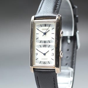 Puede incluir: Un reloj Seiko Lucent rectangular con una correa de cuero negro. La esfera del reloj está dividida en dos secciones, cada una con fondo blanco, manecillas negras y los números 12, 3, 6 y 9. El reloj tiene un marco dorado.