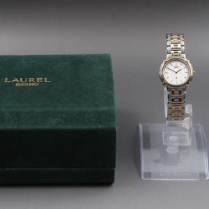 May include: A vintage Laurel Seiko watch with a silver and gold-tone band and a white face. The watch is displayed on a clear stand next to a dark green velvet box with "LAUREL SEIKO" printed in gold.