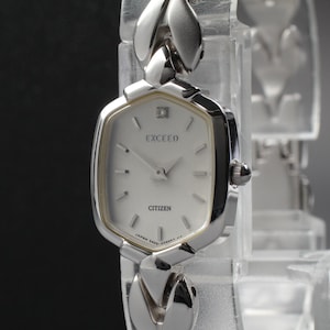 May include: A silver Citizen Exceed wristwatch with a white face and a diamond accent at the 12 o'clock position. The watch has a unique, geometric case and a linked bracelet. The watch is displayed against a dark background.