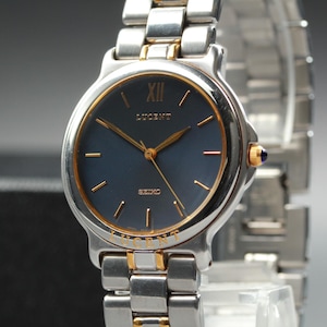 May include: A Lucent wristwatch with a silver and gold-tone stainless steel strap. The watch face is navy blue with gold hands and markers. The watch has a gold bezel and a small blue gem on the side.