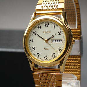 May include: Gold-toned Alba Success watch with a white face, black numerals, and hands. The watch displays the day and date, with the text "SUCCESS" and "ALBA" visible. The watch has a gold mesh band.