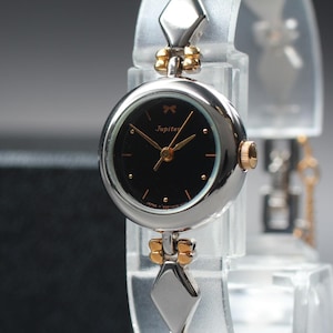 May include: A silver and gold-toned Jupiter wristwatch with a black face. The watch has gold hands and hour markers, and a small bow detail above the brand name. The band features diamond-shaped links.