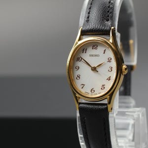 Può includere: Un orologio Seiko color oro con quadrante bianco e cinturino in pelle nera. L'orologio ha lancette e indici delle ore dorati. Il quadrante mostra il marchio Seiko. L'orologio è esposto su un supporto trasparente.