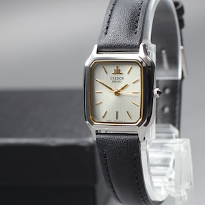Puede incluir: Un reloj Credor Seiko rectangular con caja plateada y correa de cuero negro. La esfera del reloj es clara con agujas y marcadores dorados. El reloj se exhibe sobre un soporte transparente.