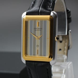 Puede incluir: Reloj Orient Quartz rectangular con bisel dorado y esfera a rayas grises y doradas. El reloj tiene agujas doradas y una correa de cuero negro. El reloj se exhibe sobre un soporte transparente.