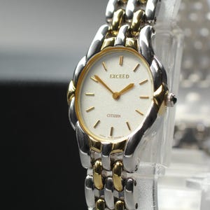May include: A Citizen Exceed wristwatch with a silver and gold-tone band and a white oval face. The watch features gold hands and markers, with the brand name "EXCEED" and "CITIZEN" displayed on the face. The watch is displayed on a clear stand.