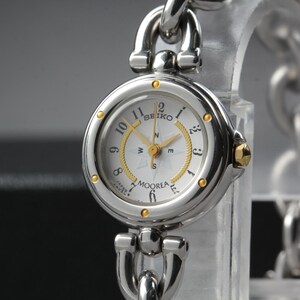 May include: A silver Seiko Moorea watch with a white face, gold accents, and a compass design. The watch has a silver chain-link band and a gold crown. The watch face displays the brand name and the word "Moorea".