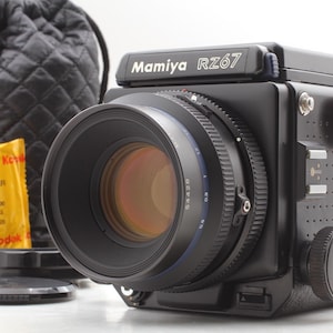May include: A black Mamiya RZ67 medium format camera with a large lens, featuring a prominent focus ring and shutter release dial. Beside the camera sits a yellow Kodak Ektar 120 film roll, a black lens cap, and a black quilted drawstring storage bag.