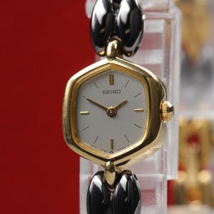 May include: A gold-toned Seiko watch with a light-colored face and black and gold-toned band. The watch face is a unique shape with gold hands and markers. The watch is displayed against a red background.