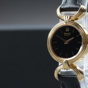 May include: A gold-tone Seiko Quartz wristwatch featuring a round black dial with a single Roman numeral at the twelve o'clock position, gold-tone hands, and a textured gold-tone bezel. The watch has a black leather strap with gold-tone lugs.