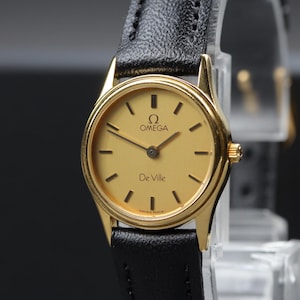 May include: An Omega De Ville wristwatch featuring a round gold-tone case, a champagne-colored dial with black hour markers, and thin black hands. The watch includes a black textured leather strap and displays the Omega logo and De Ville text.