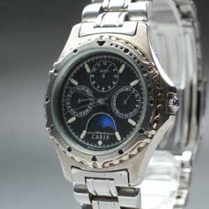May include: Silver-toned ALBA CARIB watch with a black face. The watch features multiple subdials, a moon phase indicator, and a date display. The watch has a stainless steel band and a decorative bezel.