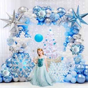 May include: A large balloon arch featuring blue, silver, and white balloons, accented with large silver and blue star-shaped foil balloons and snowflake-shaped foil balloons. A child in a light blue dress stands in front of a white Christmas tree.