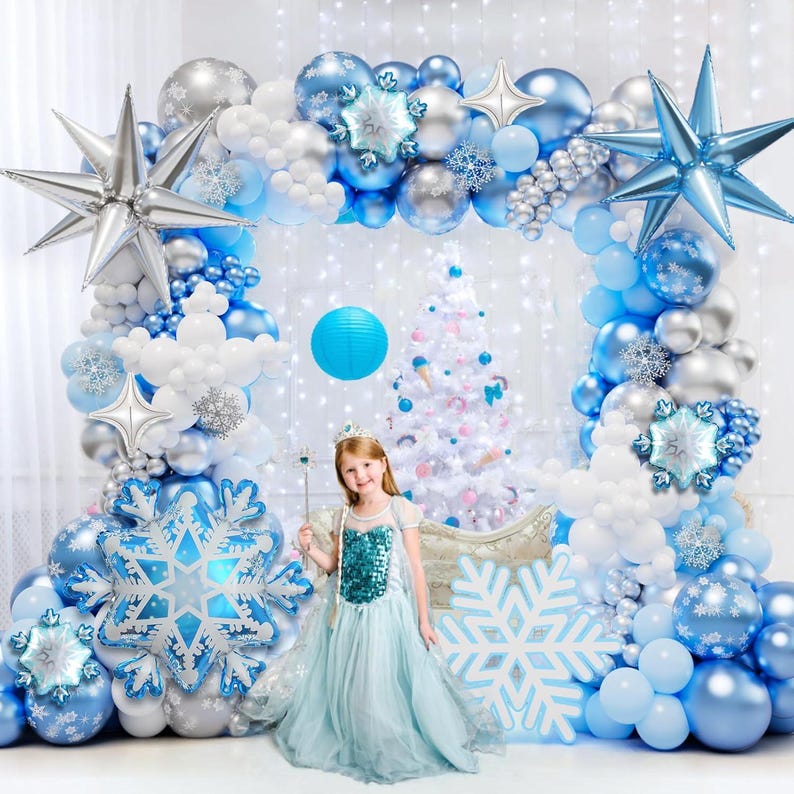 May include: A large balloon arch featuring blue, silver, and white balloons, accented with large silver and blue star-shaped foil balloons and snowflake-shaped foil balloons. A child in a light blue dress stands in front of a white Christmas tree.