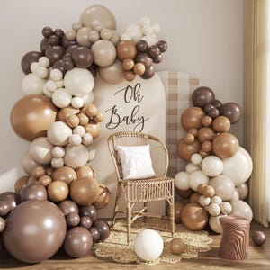 May include: A balloon arch in shades of cream, tan, and brown surrounds an arched sign with the text Oh Baby. A rattan chair with a white cushion sits in front of the sign, placed on a round woven rug with a small stool and loose balloons.