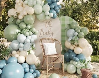 May include: A decorative balloon arch in shades of sage green, dusty blue, and cream surrounding a beige arched backdrop with the text Oh Baby. A rattan chair with a white cushion sits in front, accompanied by a small wooden stool and greenery.