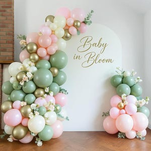 May include: A balloon arch and arrangement in pastel colors, including pink, green, gold, and white. The arch is decorated with faux flowers and greenery. The words "Baby in Bloom" are written on a white backdrop.