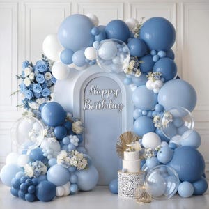 May include: A festive birthday party backdrop featuring a light blue archway adorned with a balloon garland in shades of blue and white. The words "Happy Birthday" are displayed. A cake and floral arrangements complete the celebratory scene.