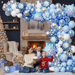 May include: A large balloon arch featuring a mix of light blue, white, and metallic silver balloons, accented with several blue and white snowflake-shaped foil balloons, creating a festive winter-themed decoration for a holiday celebration.