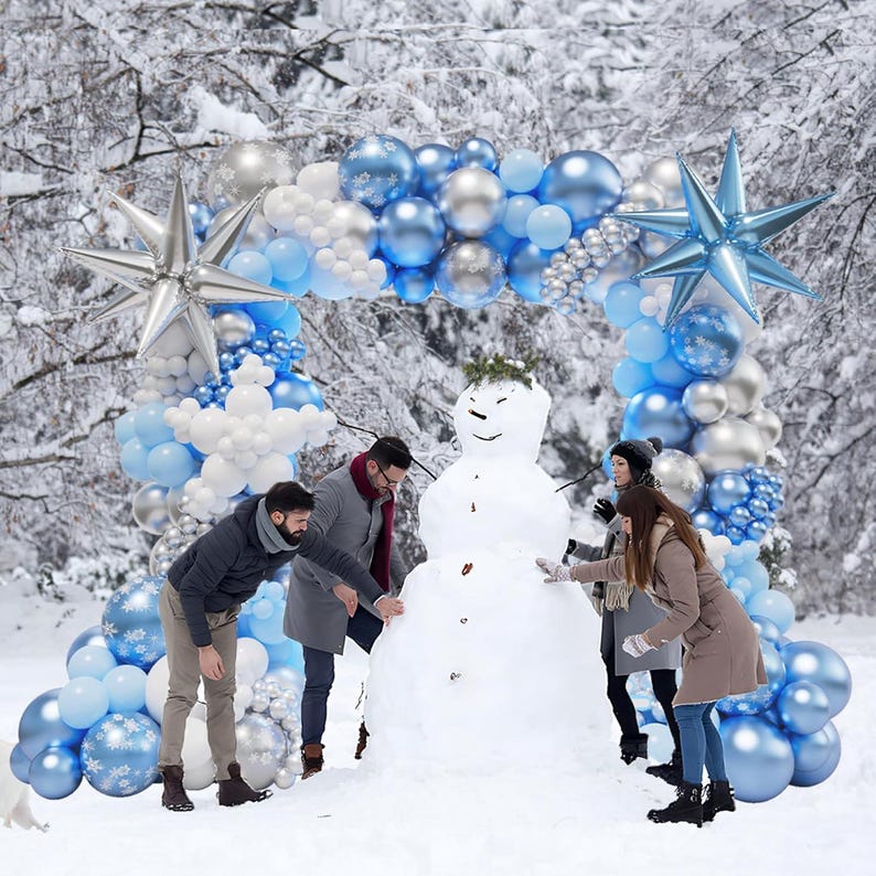 May include: A large balloon arch decoration featuring a mix of light blue, white, and metallic silver balloons, accented with large silver and blue star-shaped foil balloons, framing a snowman in a snowy outdoor setting.
