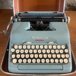May include: A vintage Smith-Corona electric typewriter in a light blue colour, with a full QWERTY keyboard and white keys. The typewriter is in its original brown case. The brand name is visible on the front.