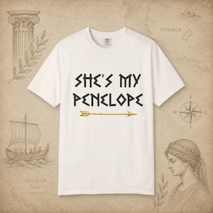 Puede incluir: Una camiseta blanca de manga corta con el texto negro "SHE'S MY PENELOPE" en una fuente estilizada, situada sobre un gráfico de flecha horizontal de color dorado. La camiseta tiene cuello redondo y un corte estándar sobre fondo neutro.
