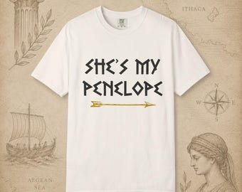 Op de afbeelding: Een wit T-shirt met korte mouwen en de zwarte tekst "SHE'S MY PENELOPE" in een gestileerd lettertype, geplaatst boven een horizontale goudkleurige pijlgrafiek. Het T-shirt heeft een ronde hals en een standaard pasvorm op een neutrale achtergrond.