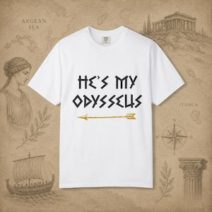 Puede incluir: Una camiseta blanca de manga corta con el texto negro HE'S MY ODYSSEUS impreso en una fuente estilizada, situado sobre un gráfico de flecha dorada horizontal. La camiseta tiene cuello redondo y un corte estándar sobre un fondo neutro.