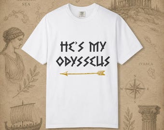 Op de afbeelding: Een wit T-shirt met korte mouwen en de zwarte tekst HE'S MY ODYSSEUS in een gestileerd lettertype, geplaatst boven een horizontale goudkleurige pijlafbeelding. Het shirt heeft een ronde hals en een standaard pasvorm op een neutrale achtergrond.