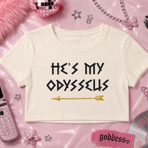 Puede incluir: Una camiseta corta de color crema con mangas cortas que presenta el texto negro "HE'S MY ODYSSEUS" impreso en la parte delantera, acompañado de un gráfico de flecha dorada colocado justo debajo del texto. La prenda tiene cuello redondo y dobladillo corto.