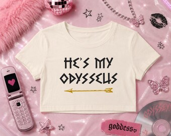 Op de afbeelding: Een crèmekleurige crop top met korte mouwen en de zwarte tekst "HE'S MY ODYSSEUS" op de voorkant, vergezeld van een gouden pijlafbeelding direct onder de tekst. Het kledingstuk heeft een ronde halslijn en een korte zoom.