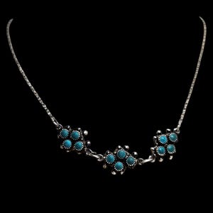 May include: A silver necklace featuring three clusters of turquoise stones. The necklace has a fine chain and the stones are set in a decorative silver setting. The background is black.