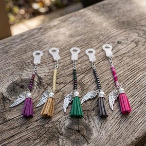 Könnte beinhalten: Fünf farbenfrohe Quastenanhänger mit silbernen Engelsflügel-Akzenten. Jeder Anhänger hat eine andersfarbige Quaste: lila, beige, grün, grau und rosa. Die Anhänger sind mit Perlenketten an transparenten Kunststoffschlaufen befestigt.